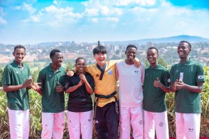 Eine Gruppe junger Taekwondo-Sportlerinnen und -Sportler steht lachend nebeneinander im Freien; in der Mitte ein Trainer im goldfarbenen Dobok.