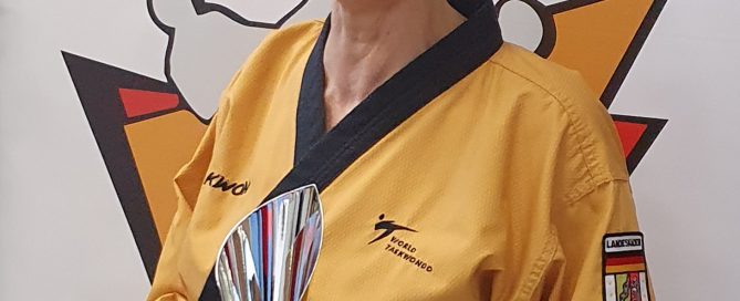 Eine Frau im goldfarbenen Taekwondo-Dobok hält einen Pokal und lächelt in die Kamera.