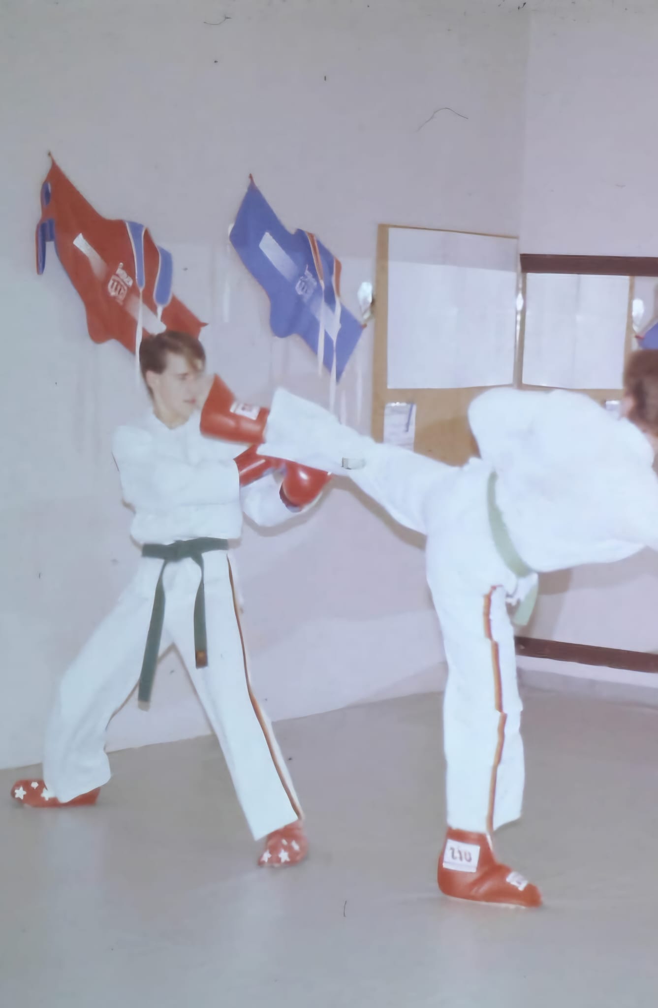 Zwei junge Taekwondo-Sportler in Schutzausrüstung trainieren in einer Halle. Einer führt einen hohen Rückwärtskick aus, der andere blockt mit roten Handschuhen. Im Hintergrund hängen rote und blaue Wettkampfweste-Sets an der Wand. Aufnahme von etwa 1985.