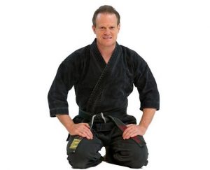Person in schwarzem Kampfkunstanzug sitzt im Kniesitz und lächelt in die Kamera.