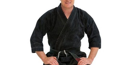 Person in schwarzem Kampfkunstanzug sitzt im Kniesitz und lächelt in die Kamera.