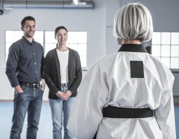 Taekwondo-Trainerin im Dobok spricht mit zwei lächelnden Erwachsenen in einem Trainingsraum.