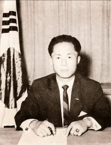 Schwarzweißfoto eines Mannes im Anzug, der am Schreibtisch sitzt und schreibt.