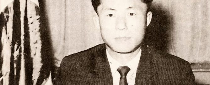 Schwarzweißfoto eines Mannes im Anzug, der am Schreibtisch sitzt und schreibt.