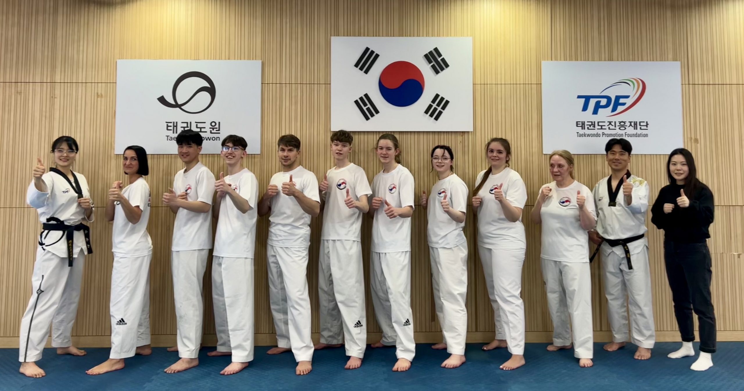 Eine Gruppe Taekwondo-Sportlerinnen und -Sportler steht in einer Linie und zeigt mit Daumen nach oben in die Kamera.