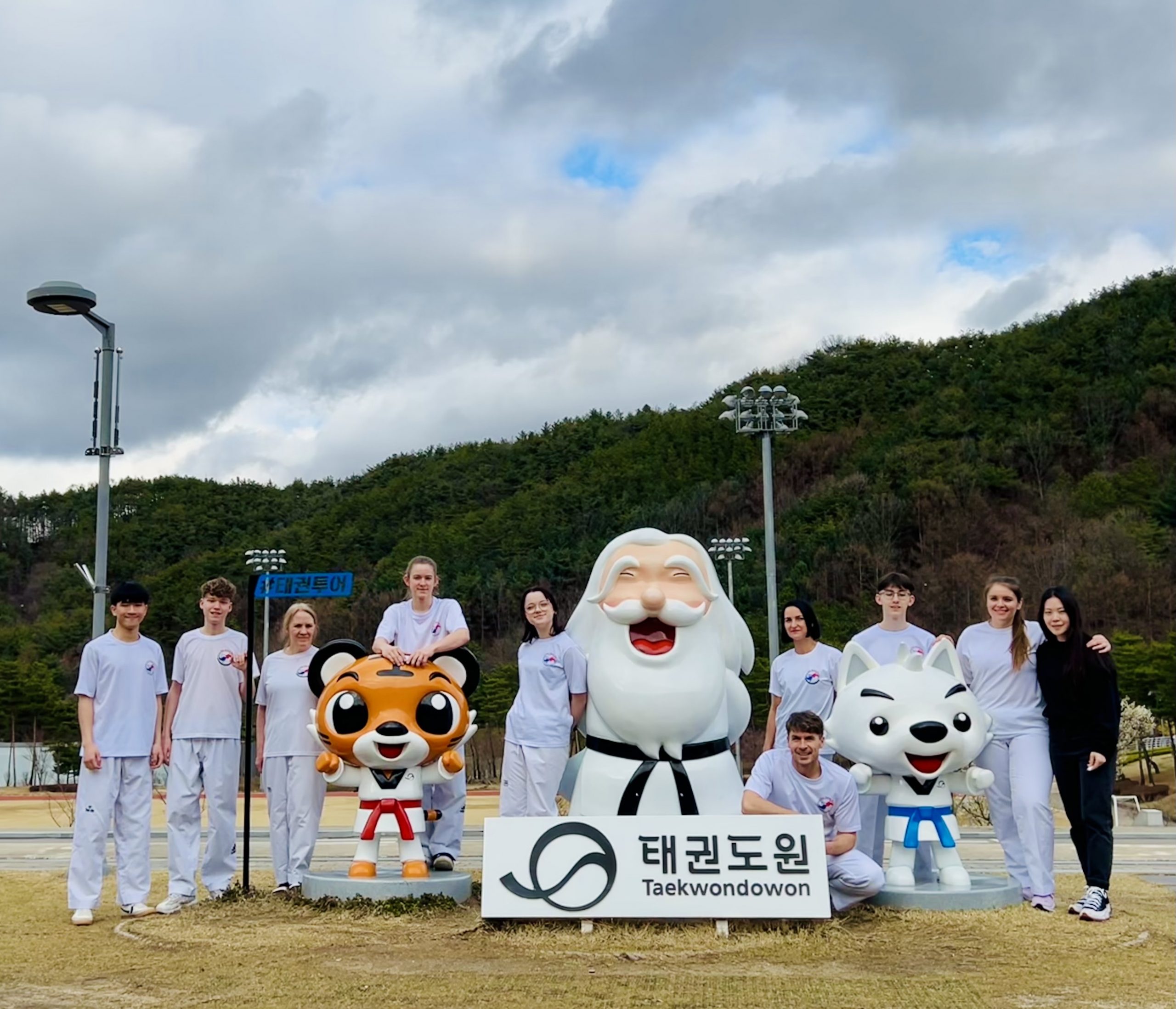 Eine Gruppe junger Taekwondo-Sportlerinnen und -Sportler posiert im Taekwondowon neben drei großen Maskottchenfiguren.