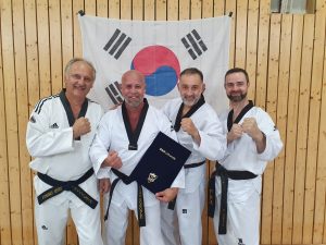 Vier Taekwondo-Sportler in weißen Doboks mit schwarzen Gürteln posieren lächelnd vor einer südkoreanischen Flagge. Einer hält eine Urkunde.