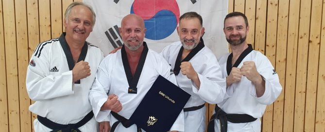 Vier Taekwondo-Sportler in weißen Doboks mit schwarzen Gürteln posieren lächelnd vor einer südkoreanischen Flagge. Einer hält eine Urkunde.