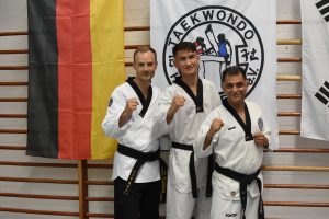 Drei Taekwondo-Meister in Doboks posieren mit erhobener Faust vor deutschen und koreanischen Flaggen.