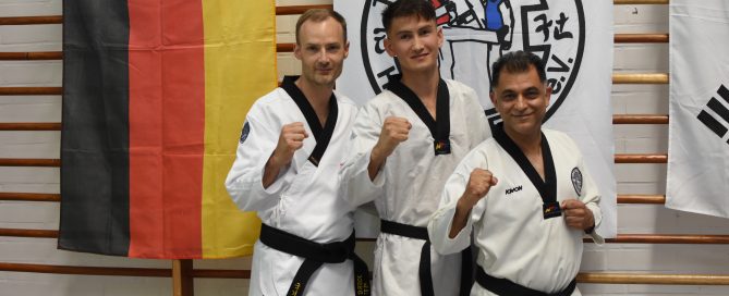 Drei Taekwondo-Meister in Doboks posieren mit erhobener Faust vor deutschen und koreanischen Flaggen.