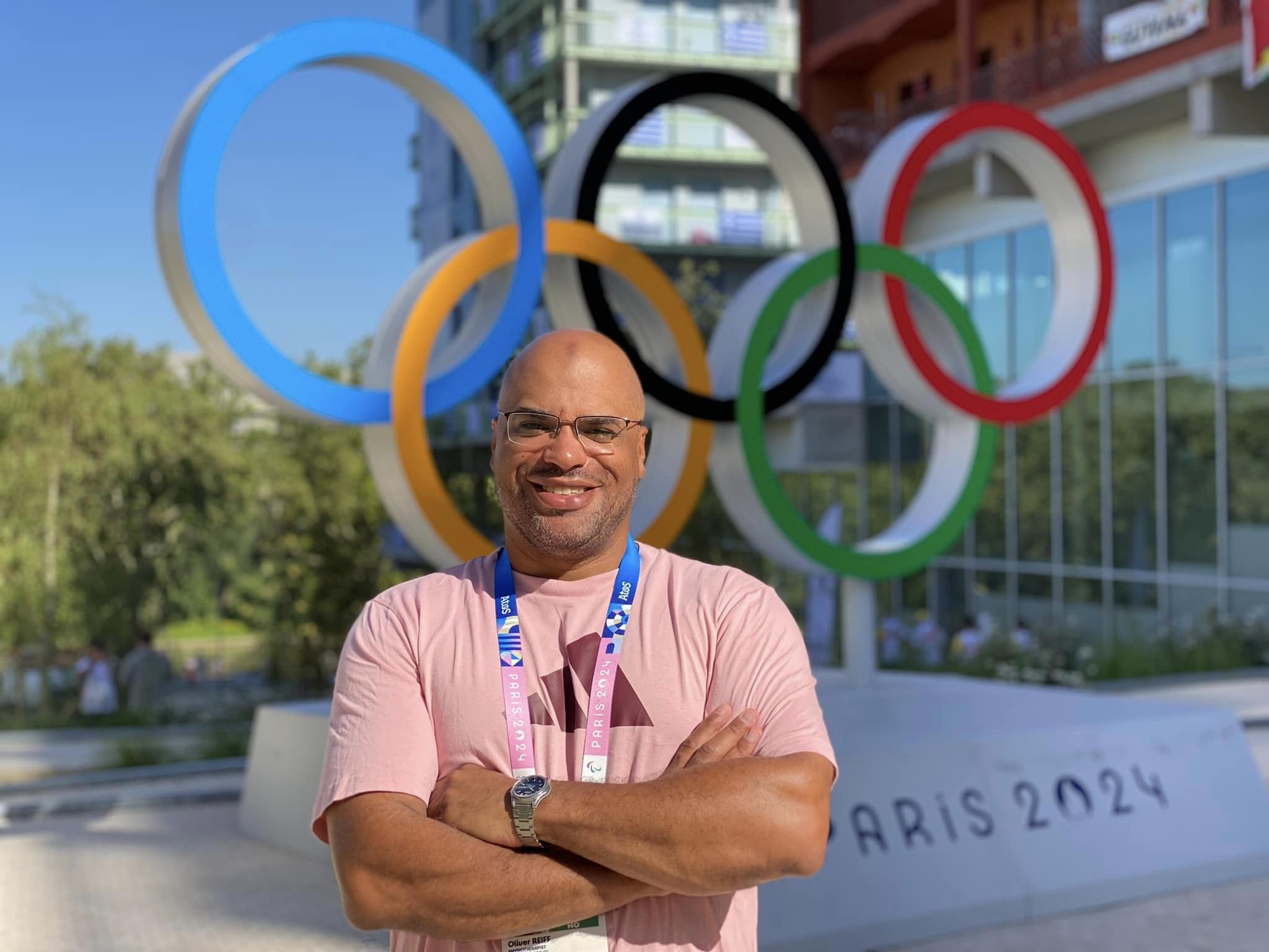 Ein Mann steht lächelnd vor den großen olympischen Ringen und trägt eine Paris-2024-Akkreditierung um den Hals.