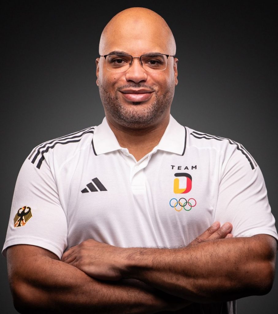 Portrait_1 Ein Mann in einem weißen Team-D-Shirt steht mit verschränkten Armen vor dunklem Hintergrund und lächelt leicht.