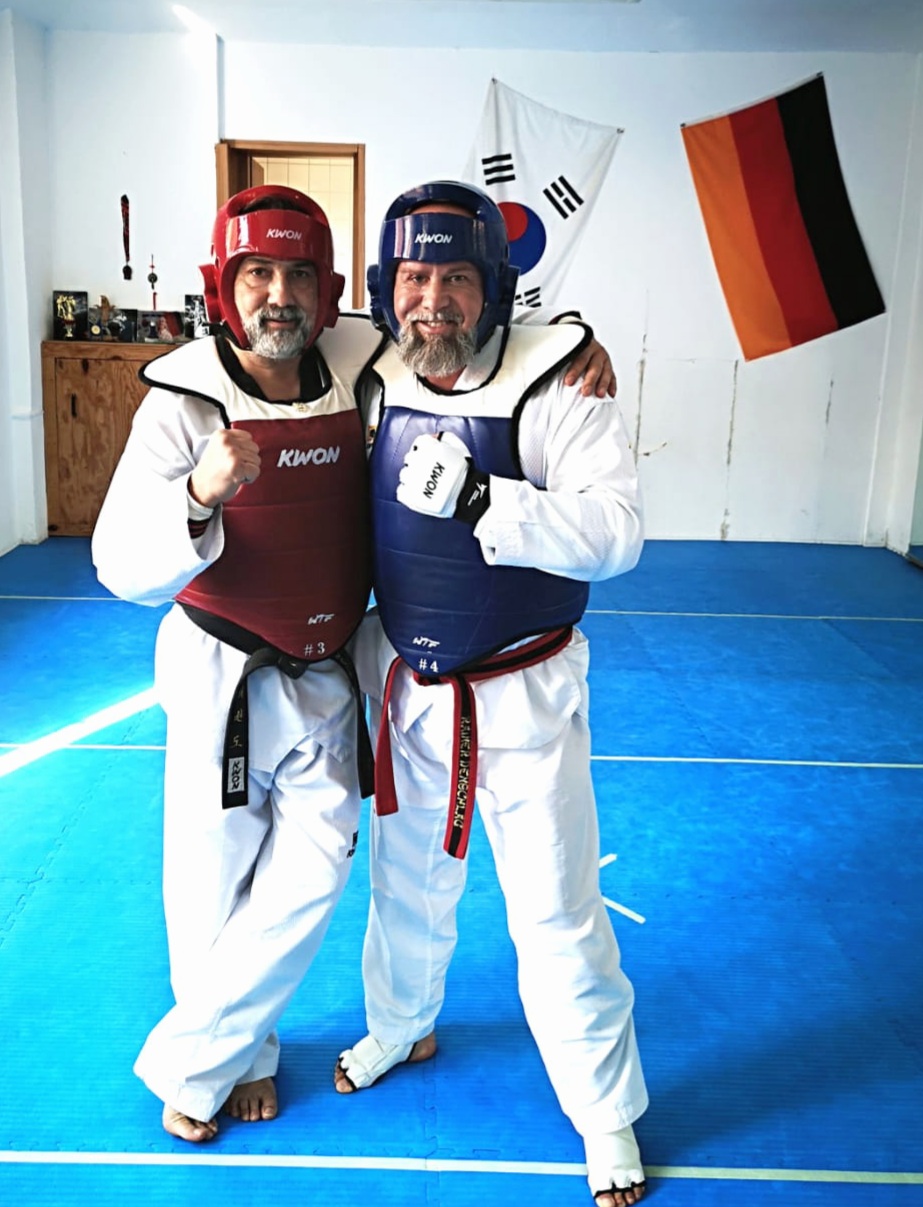 Zwei Taekwondo-Sportler, Rainer Denschlag (rechts), in Schutzwesten stehen Arm in Arm auf der blauen Matte.
