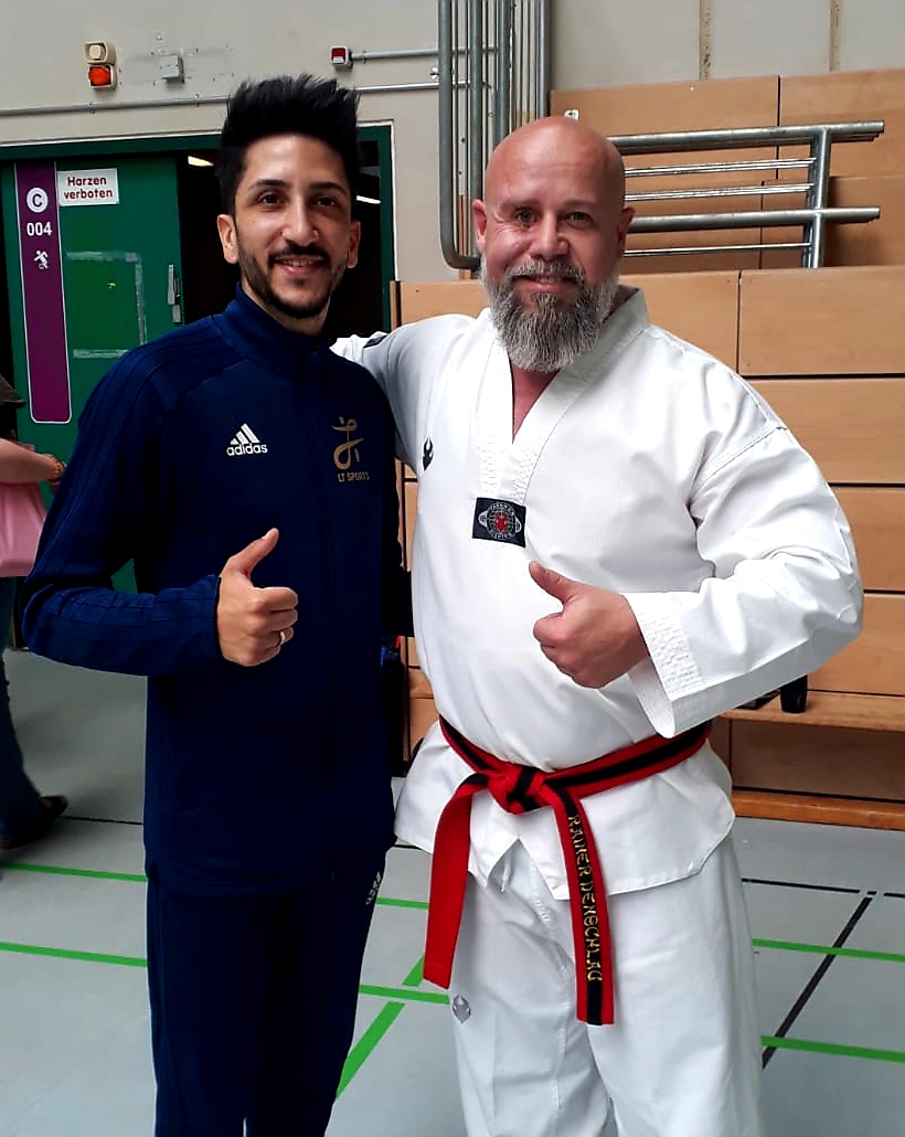 Zwei Männer stehen in einer Sporthalle nebeneinander und zeigen den Daumen nach oben. Rechts im Bild steht Rainer Denschlag in Taekwondo-Dobok mit rot-schwarzem Gürtel; links steht ein Mann in dunkelblauer Trainingskleidung.