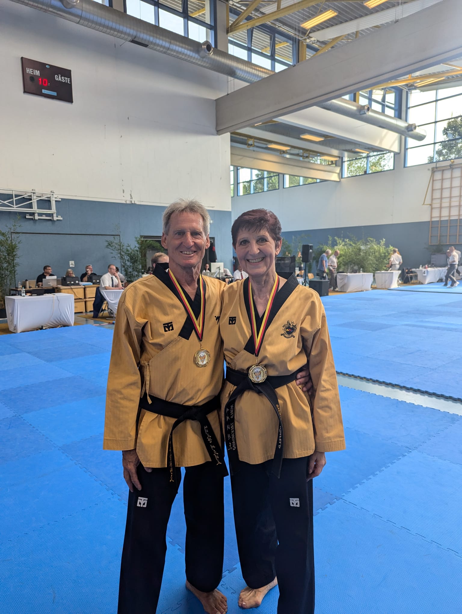 Zwei Taekwondo-Sportler im gold-schwarzen Dobok stehen in einer Sporthalle und tragen Medaillen.