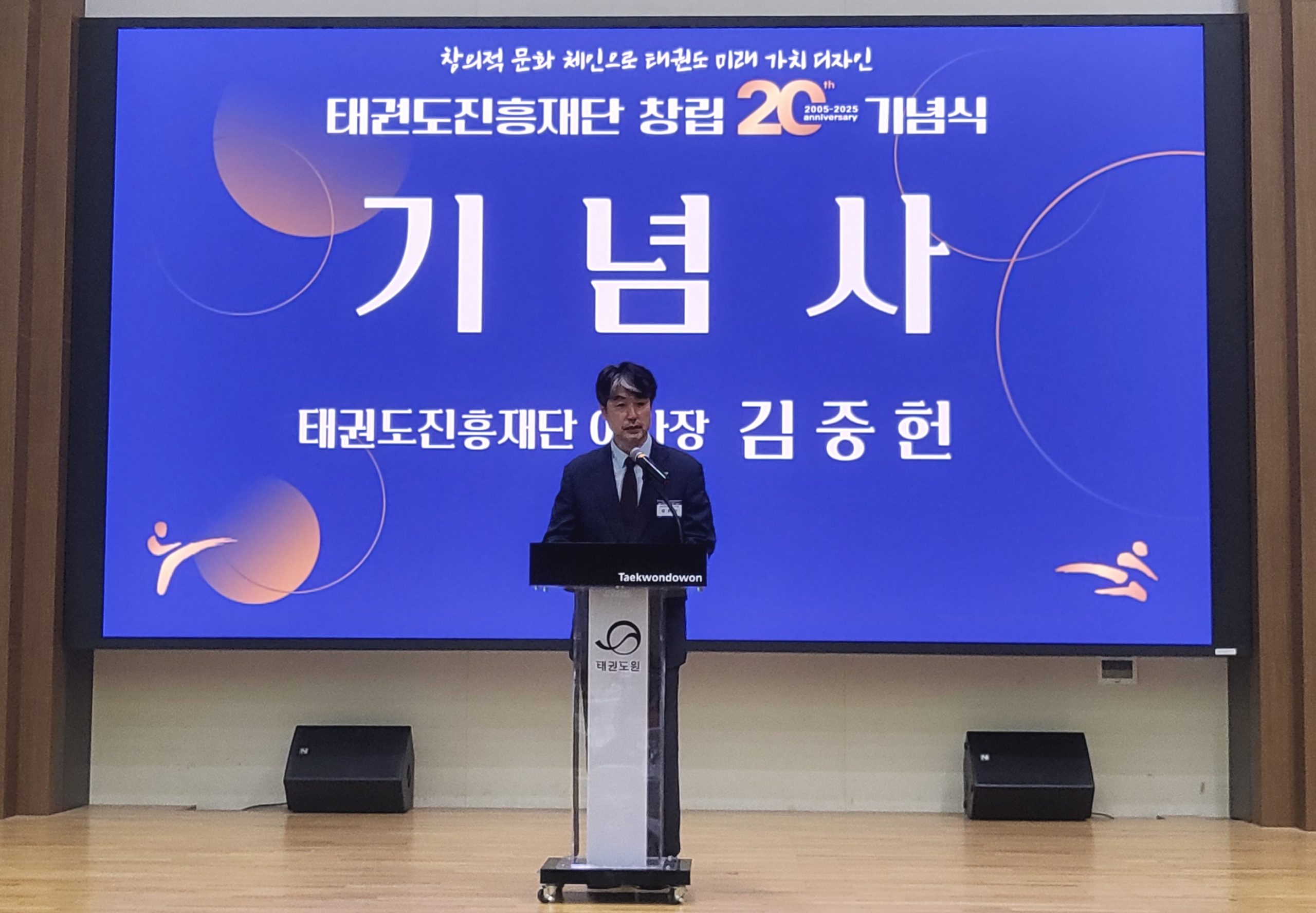Ein Mann steht am Rednerpult auf einer Bühne vor einer großen blauen Präsentationswand mit koreanischer Schrift.