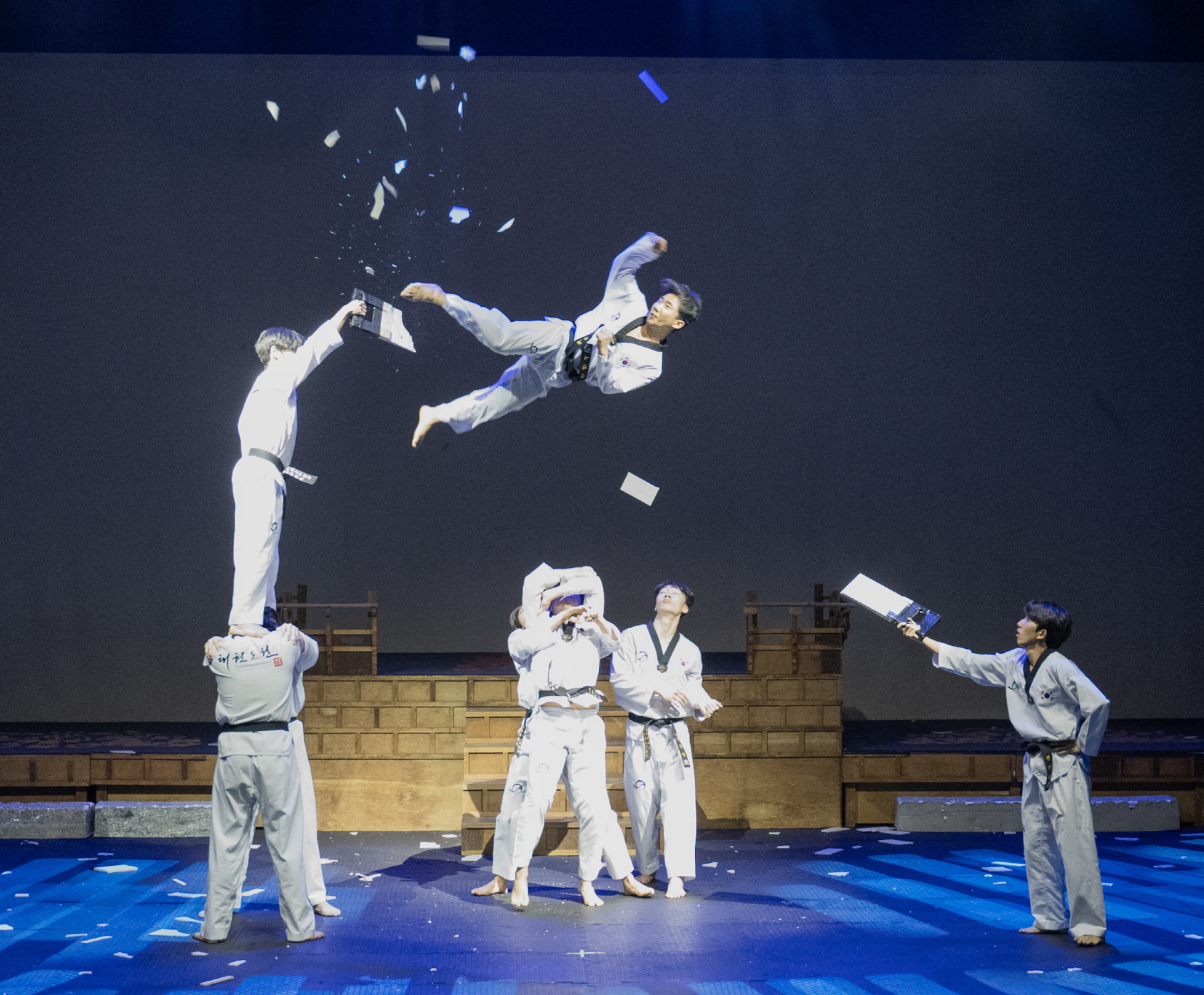 Ein Taekwondo-Demonstrationssportler springt in der Luft und zerbricht ein Holzbrett, während Teammitglieder um ihn herum stehen.