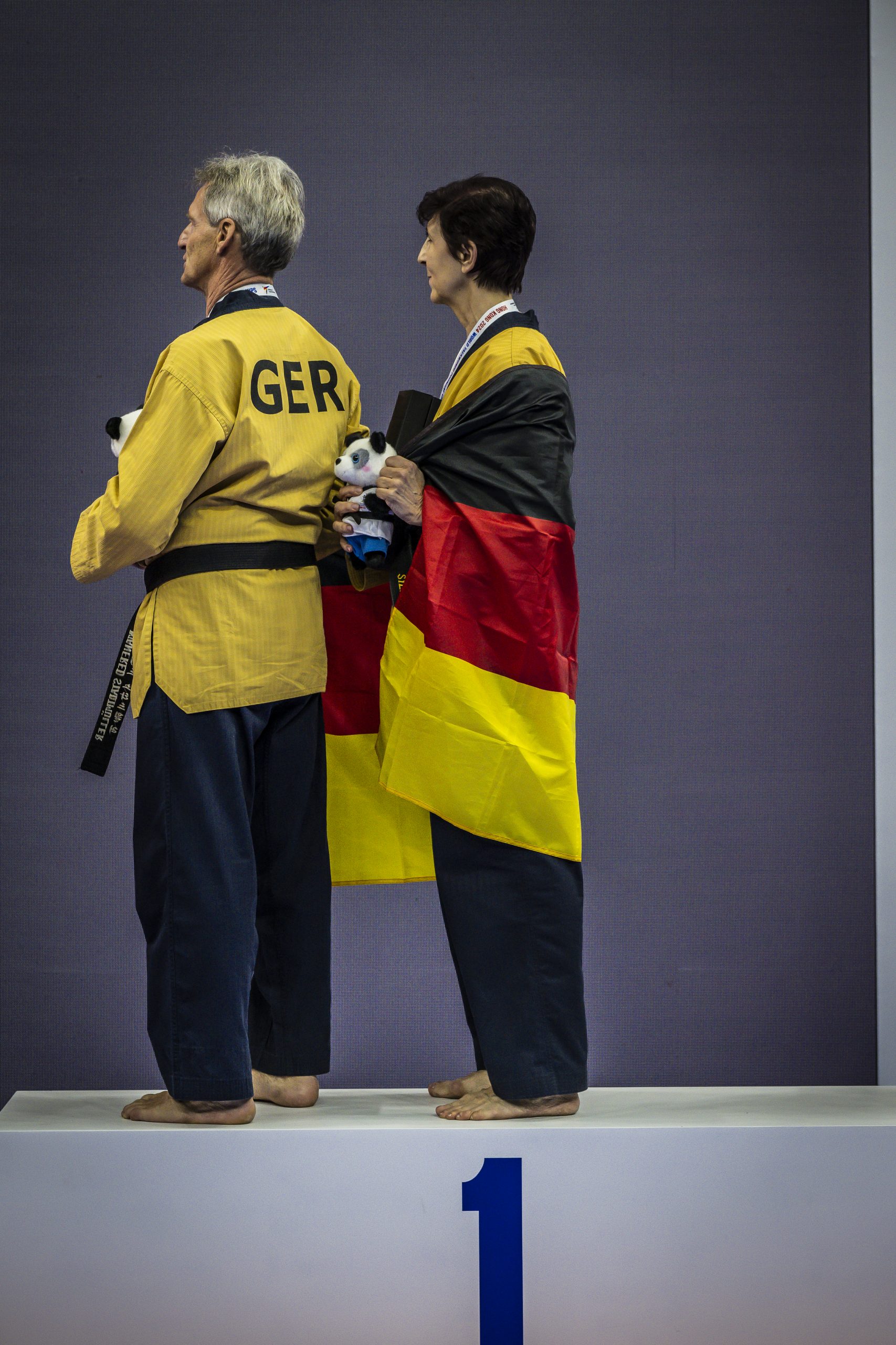 Zwei Taekwondo-Sportler stehen auf dem Siegerpodest, in goldenen Doboks und mit einer Deutschlandflagge umgehüllt.