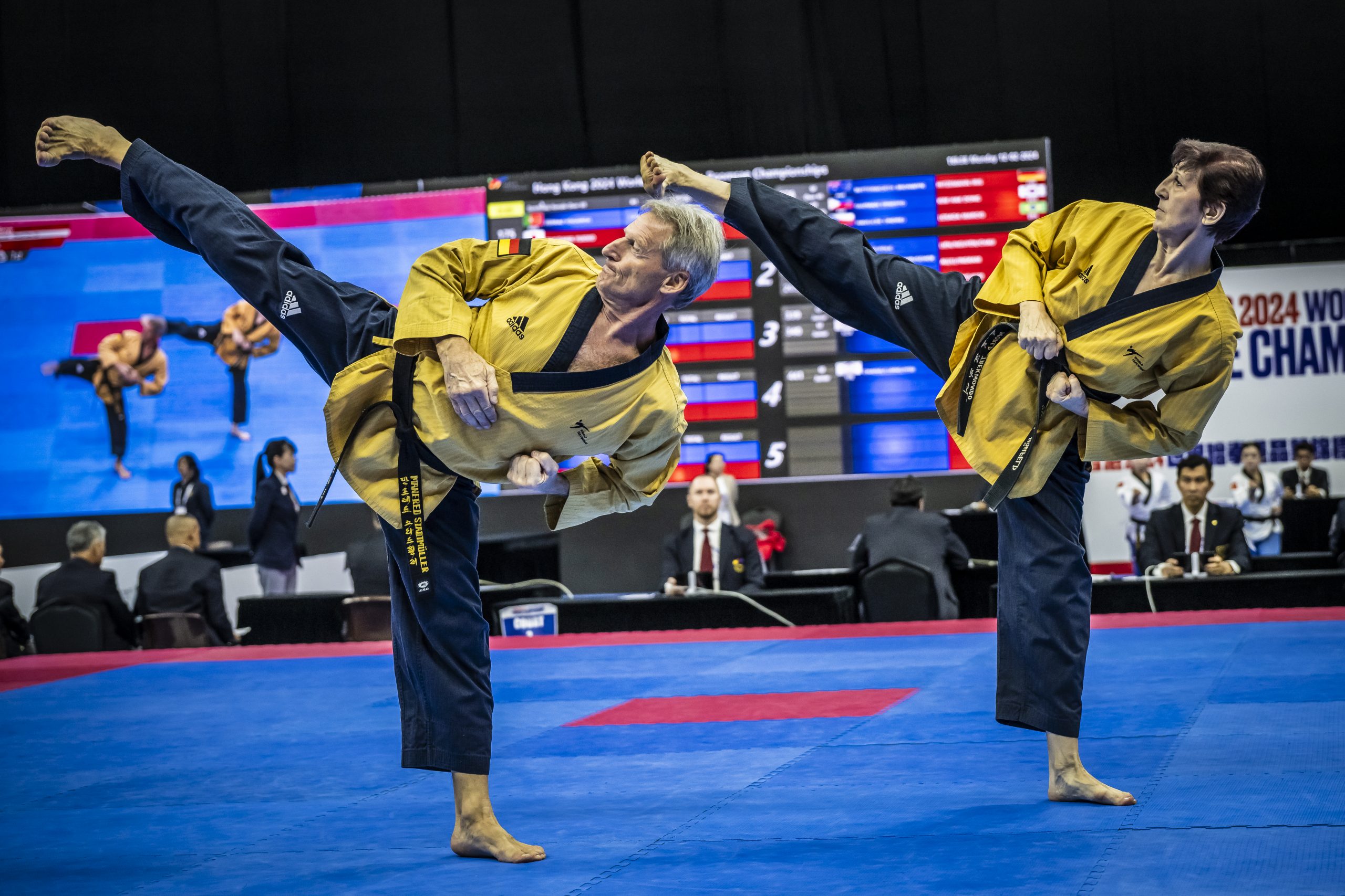 Zwei Taekwondo-Sportler führen auf der Wettkampfmatte synchron hohe Seittritte aus.