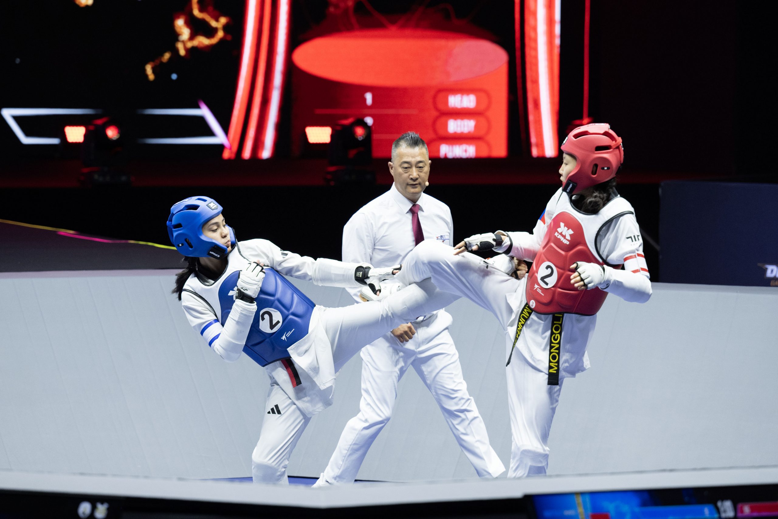 Zwei Taekwondo-Kämpferinnen treten im Wettkampf gegeneinander an; eine trägt blaue, die andere rote Schutzausrüstung. Ein Kampfrichter beobachtet die Aktion.